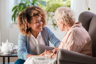 Female home caregiver talking with senior woman, sitting in living room and listening to her carefully.
