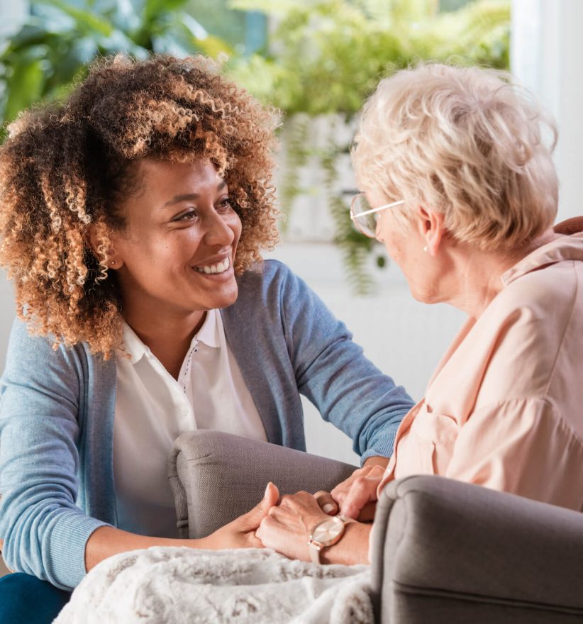 Female home caregiver talking with senior woman, sitting in living room and listening to her carefully.